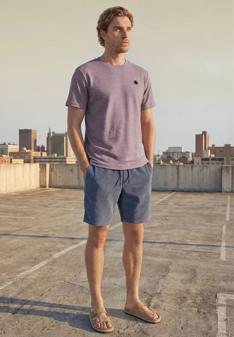 Ein Mann steht auf einem Dach mit Stadtblick, trägt lila T-Shirt, blaue Shorts und Sandalen.