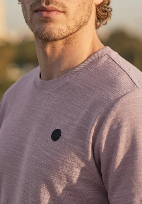 Ein junger Mann in einem dezenten, fliederfarbenen Pullover mit kleinem, schwarzem Logo vor unscharfem Hintergrund.