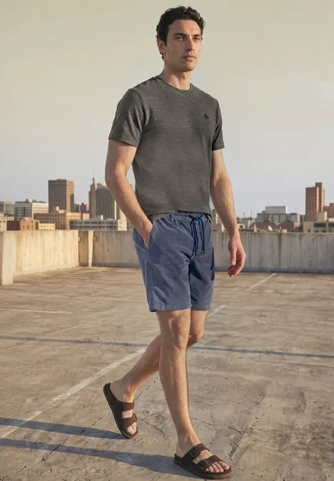Ein Mann steht auf einem Parkdach, trägt graues T-Shirt, blaue Shorts und Sandalen, Skyline im Hintergrund.