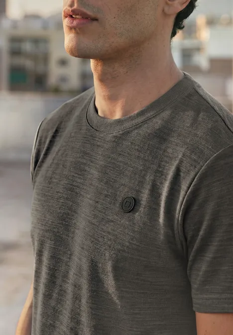 Ein Mann trägt ein graues T-Shirt mit kleinem Brustlogo, unscharfer urbaner Hintergrund.