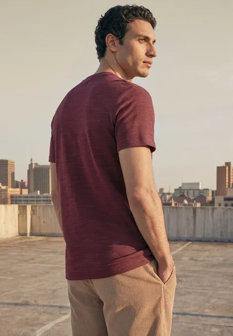 Ein Mann steht auf einem Dach, blickt über die Stadt und trägt ein rotbraunes T-Shirt mit beigefarbener Hose.