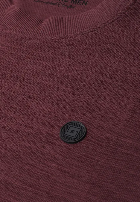 Bordeauxfarbenes gestricktes T-Shirt mit schwarzem rundem Logoemblem auf der Brust.