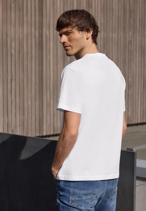 Een jonge man staand in profiel voor een houten wand, met een wit T-shirt en een blauwe spijkerbroek.