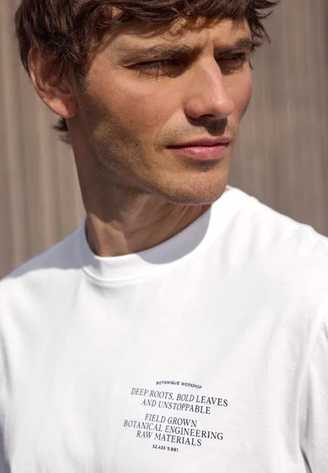 Portret van een man in een wit T-shirt met tekst, voor een houten wand.