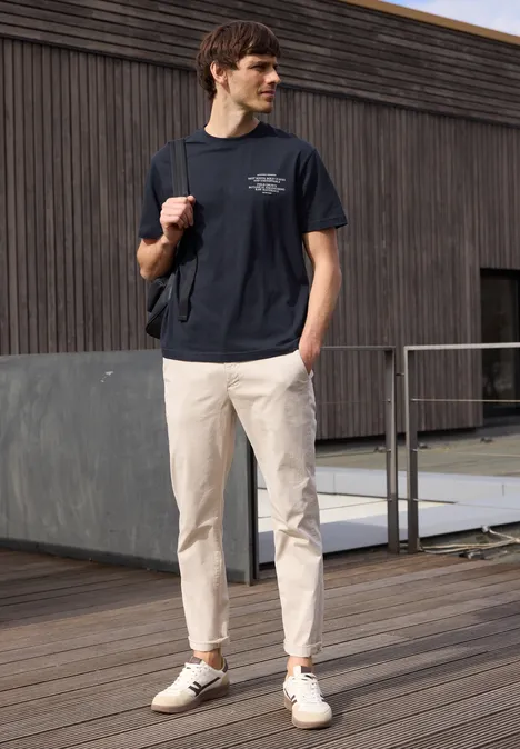 Een man op een houten terras draagt een donker T-shirt, lichte chino’s en witte sneakers, met een rugzak over één schouder.