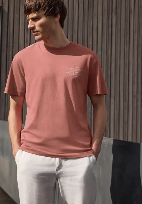 Man in roze t-shirt en witte broek voor een houten wand.