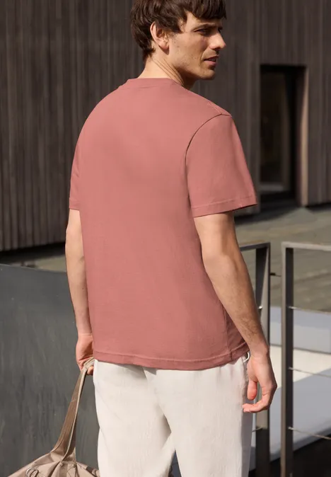 Een man van achteren in een roze T-shirt en lichte broek, met een tas voor een moderne houten gevel.