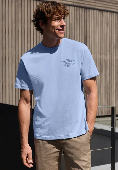 Ein Mann steht in hellblauem T-Shirt und beiger Hose vor einer Holzfassade.