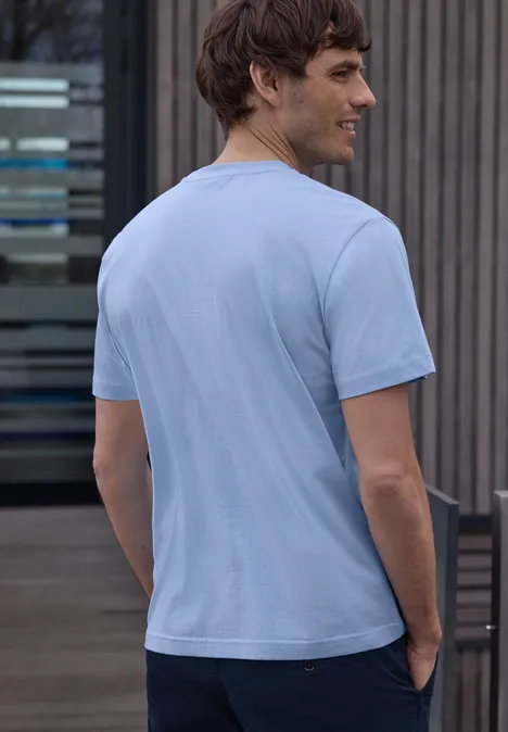 Een man staat met zijn rug naar de camera, draagt een lichtblauw T-shirt en een donkere broek voor een modern gebouw.