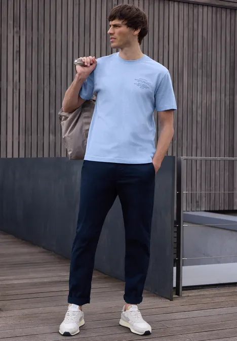 Een man in een casual outfit met lichtblauw T-shirt, donkerblauwe broek en witte sneakers buiten.