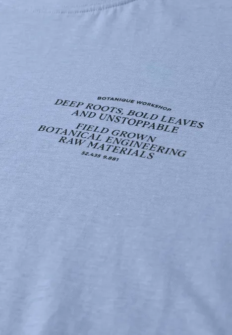 Lichtblauw T-shirt met zwarte botanische tekst over wortels, bladeren en opbrengsten.