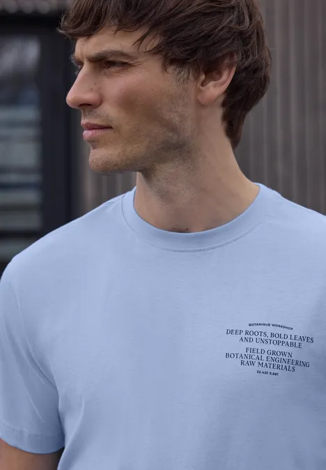 Een zijdelings afgebeelde man draagt een lichtblauw t-shirt met klein zwart logo, onscherp gezicht.