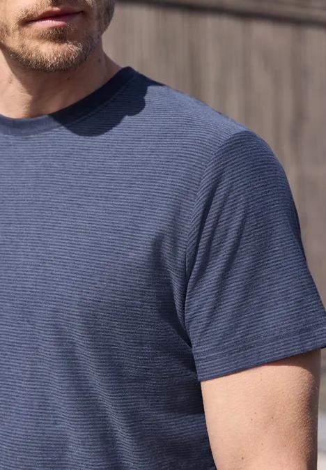 Een man met lichte baard draagt een licht gestreept blauw T-shirt voor een houten wand.