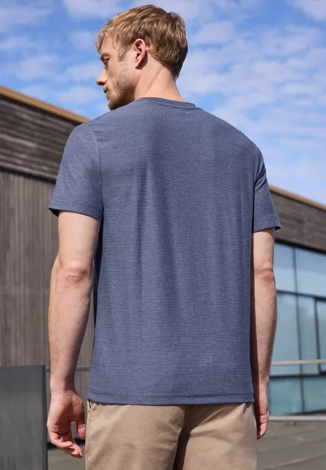 Een man in een blauw T-shirt en beige broek gezien van achteren voor een modern gebouw en blauwe lucht.