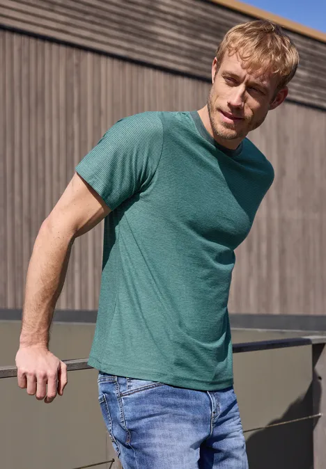 Een man in een groen T-shirt en spijkerbroek leunt nonchalant tegen een balustrade voor een houten muur.