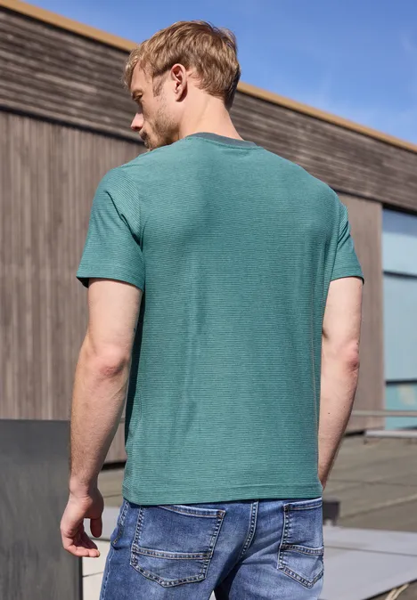 Man van achteren in groen T-shirt en blauwe spijkerbroek voor een houten gevel bij blauwe lucht.
