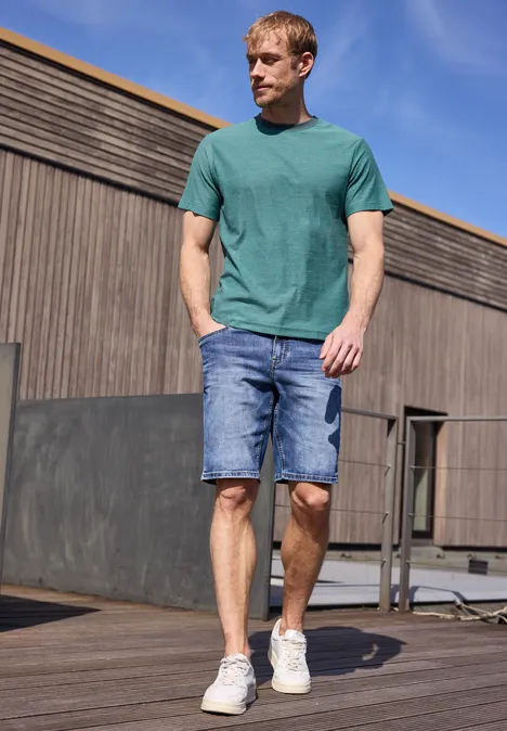 Een man in een groen t-shirt en spijkershort loopt op een houten vlonder voor een modern gebouw.