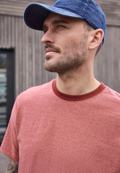 Ein Mann mit rotem T-Shirt und blauer Baseballkappe vor einer Holzwand.