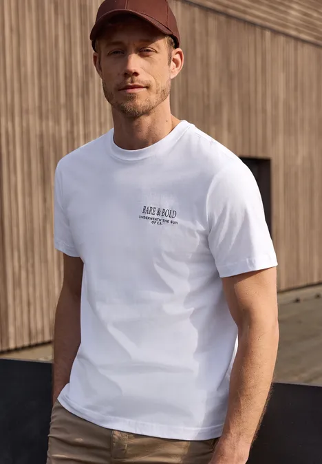 Man in wit t-shirt en rode pet voor een houten gevel.