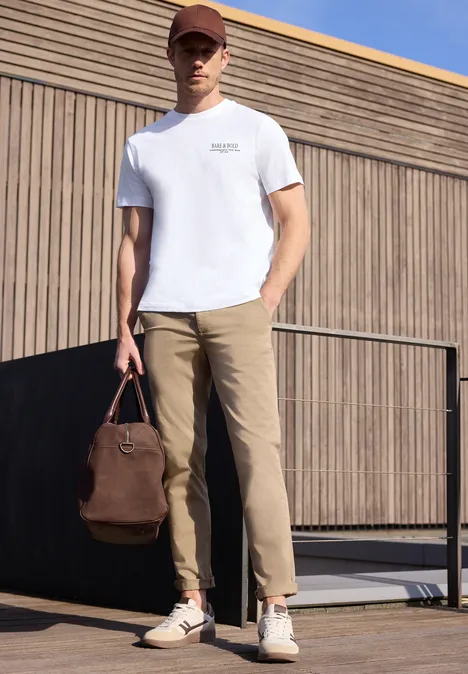 Een man in wit T-shirt en beige broek houdt een bruine tas vast voor een modern gebouw.
