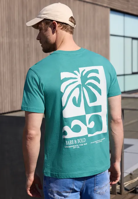 Een man van achteren in spijkerbroek en pet, met een turquoise T-shirt met witte palm- en golfprint.