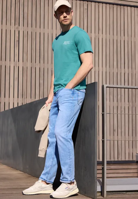Een man in een turquoise T-shirt, jeans en witte sneakers leunt ontspannen tegen een donkere balustrade voor een houten wand.