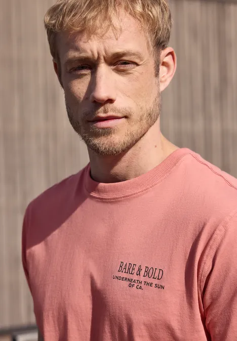 Een man draagt een roze sweatshirt met de tekst 'RARE & BOLD' voor een houten achtergrond.