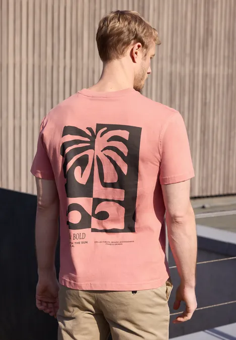 Een jonge man staat buiten en draagt een roze t-shirt met een zwarte print van palmbomen en zonnebrillen.