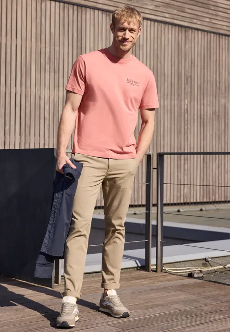 Jonge man in rood T-shirt en beige broek houdt jas vast op een terras voor een houten gevel.