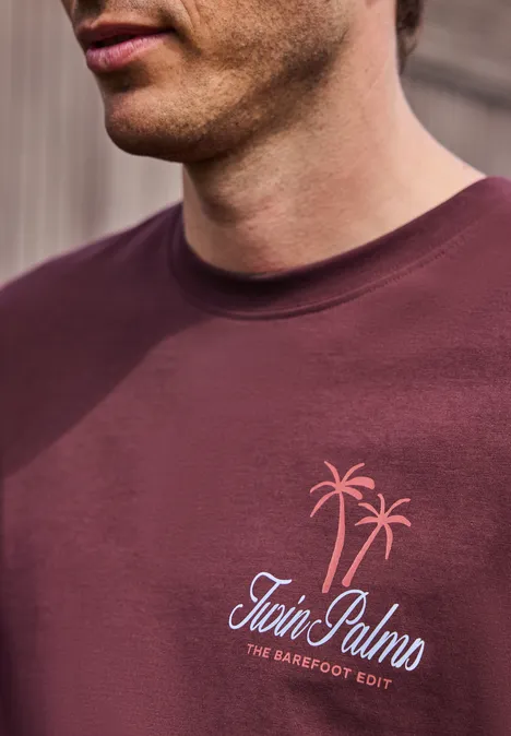 Nahaufnahme eines Mannes in bordeauxrotem T-Shirt mit Palmenmotiv und Schriftzug „Twin Palms“