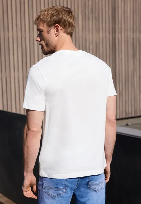 Een man in wit T-shirt en spijkerbroek staat met zijn rug naar de camera voor een houten gevel.