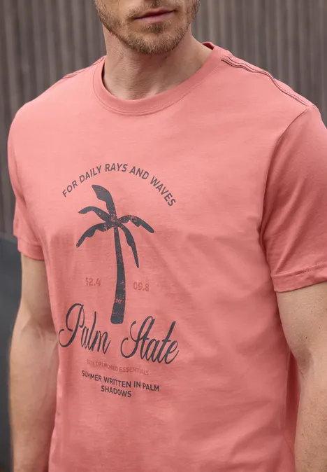 Een man draagt een zalmkleurig T-shirt met een palmbomenprint en de tekst “Palm State”.