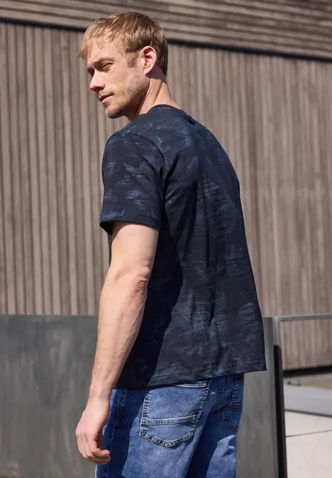 Een man in een zwart T-shirt en blauwe spijkerbroek staat voor een houten gevel.