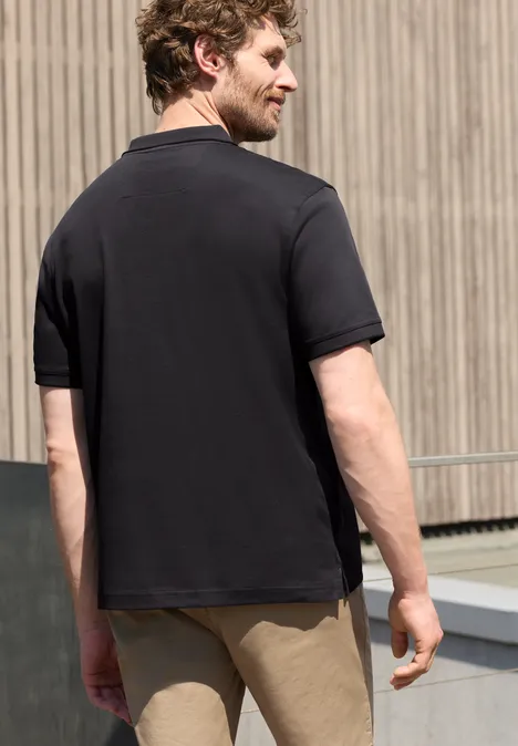 Un homme de dos porte un polo noir et un short beige devant une façade en bois.