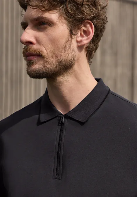 Un homme porte un polo noir à fermeture éclair, visage flou, devant un fond neutre.
