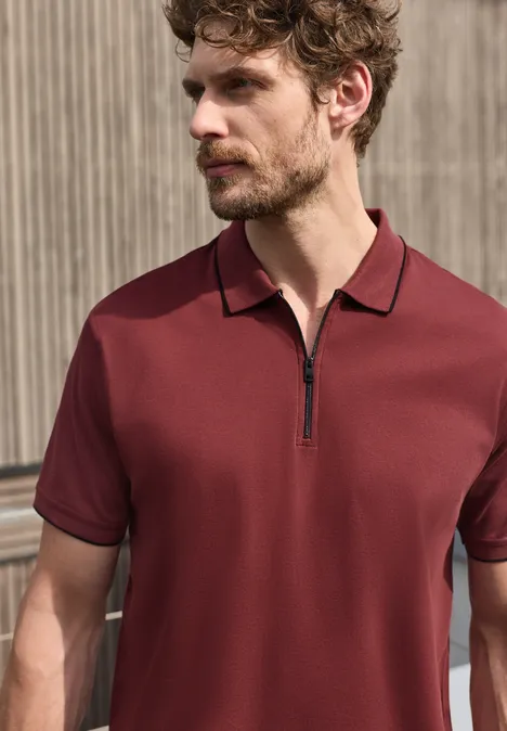Un homme porte un polo bordeaux à manches courtes avec fermeture éclair partielle.