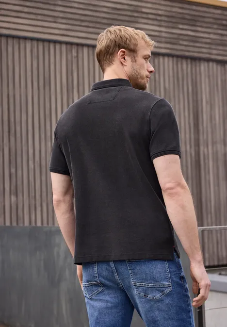 Een man van achteren in een zwart T-shirt en spijkerbroek voor een houten muur