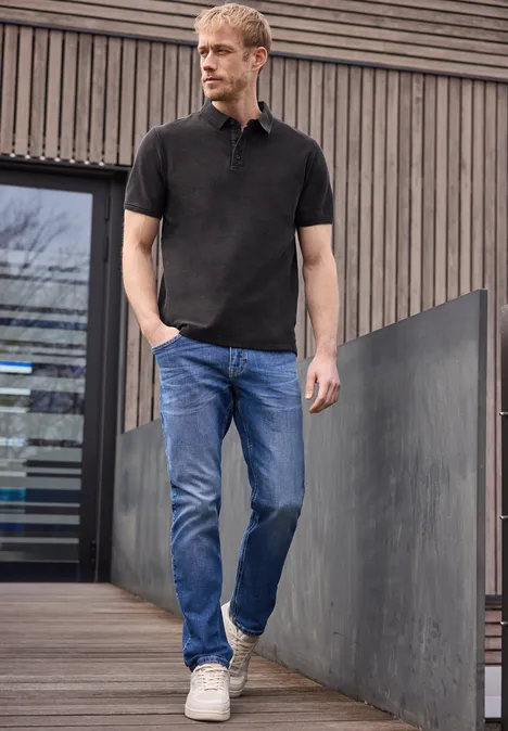 Een man staat voor een modern houten gebouw, draagt een zwart poloshirt, blauwe jeans en witte sneakers.
