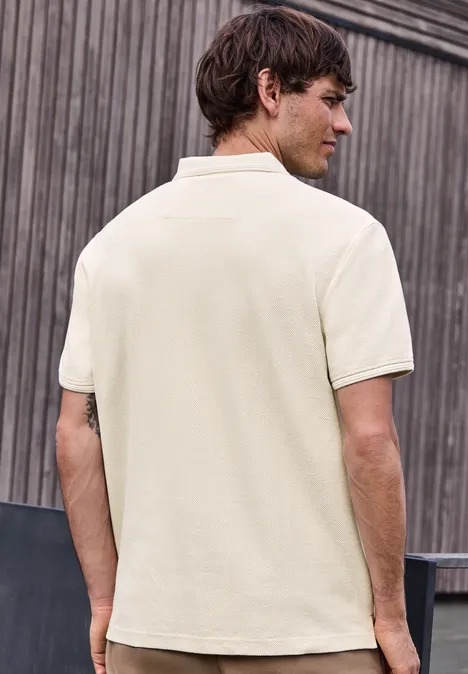Ein Mann von hinten in einem beigen Poloshirt vor einer Holzverkleidung.