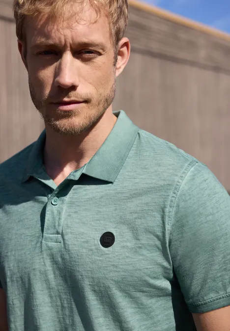 Een man in een groen poloshirt staat voor een houten wand, gezicht wazig gemaakt.