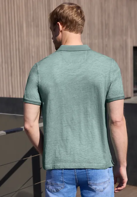 Een man met de rug naar de camera draagt een lichtgroene polo en blauwe jeans op een balkon.