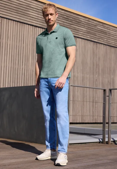 Een man in een polo en spijkerbroek staat voor een houten gevel buiten.