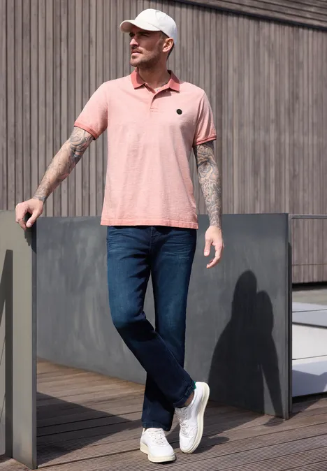 Ein Mann mit Tattoos trägt rosa Polo, Jeans und weiße Sneaker, lehnt lässig an einer Holzfassade.