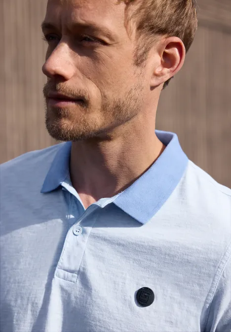 Close-up van een man met kort haar en lichtblauw poloshirt, kijkend opzij voor een houten wand.