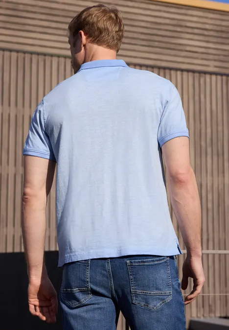 Een man gezien van achteren draagt een lichtblauw poloshirt en jeans voor een houten gevel.