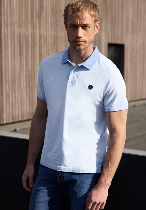 Een man draagt een lichtblauw poloshirt en spijkerbroek, staand voor een houten gevel in de buitenlucht.