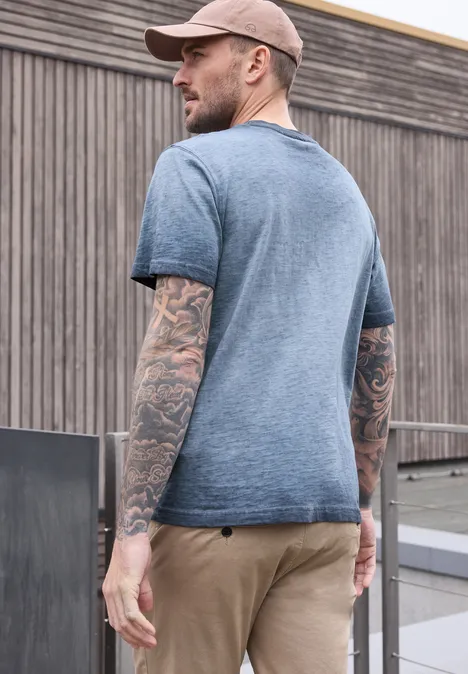 Ein Mann mit Baseballcap, Tattoos auf den Armen, trägt graues T-Shirt und steht vor einer Holzfassade.