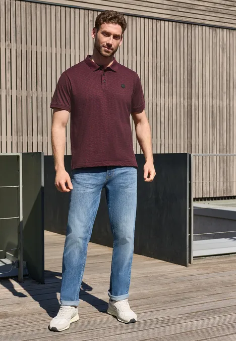 Mann in Poloshirt und Jeans steht auf Holzterrasse vor modernem Gebäude.