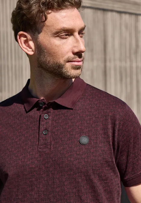 Ein Mann in einem dunkelroten Poloshirt mit Logo, vor einer hellen Holzpaneelwand.