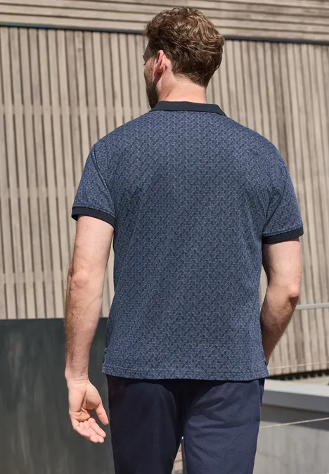 Mann von hinten trägt ein blau gemustertes T-Shirt und dunkelblaue Shorts vor einer Holzfassade.